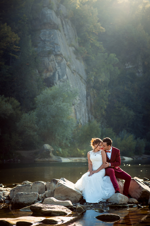 Vertical portrait of the sensitive hugging newlywed couple touching noses and resting on the stone near the river.の写真素材