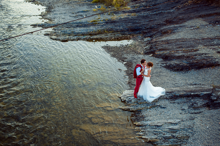 Romantic outdoor wedding shot of the fashionable newlywed couple hugging at the river bank. Beautiful landscape.の写真素材