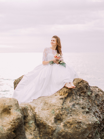 The bride in the long wedding dress is sitting on the cliff and watching the sunset at the background of the sea.の写真素材