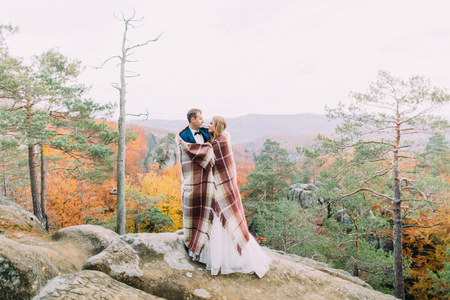 The horizontal photo of the newlywed couple wrapped in the knitted plaid standing on the rocks.の写真素材