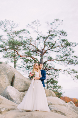 Full-length photo of the groom hugging the bride back while standing on the rock.の写真素材
