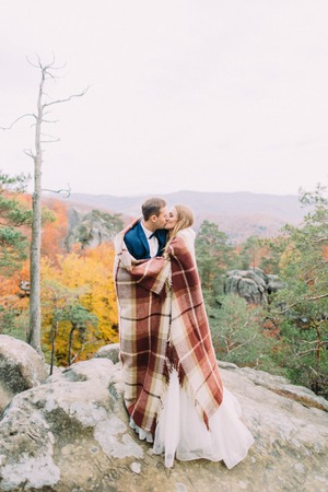Full-length photo of the kissing newlywed couple wrapped in the knitted plaid. Mountains location.の写真素材