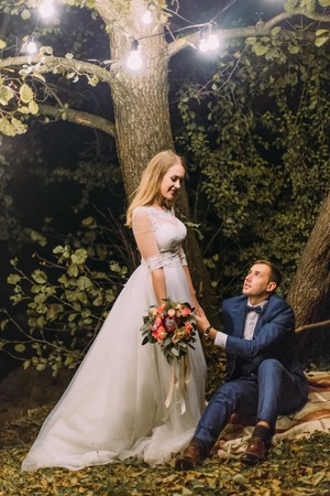 Lovely portrait of the groom holding the hand of the bride below the tree decorated with garland lamps.の写真素材