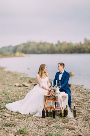 Happy newlyweds are holding hands while sitting at the wedding table on the coast. Vertical photo.の写真素材
