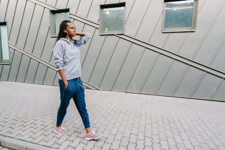 Full-length view of the young confident african girl in sportswear walking along the street and enjoying music in earphones.の写真素材