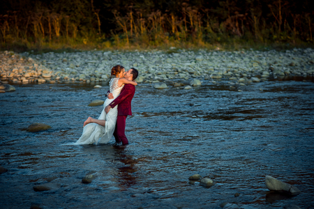 Horizontal wedding shot of the beautiful newlyweds. The groom in red suit is kissing and lifting up his charming bride while standing in the middle of the river during the sunset.の写真素材