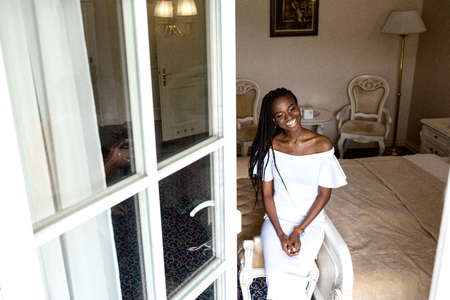 Positive beautiful afro american woman sitting in light big room in white dress in fronf of window. Positive human emotionsの写真素材