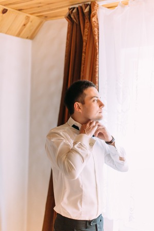 Half-length portrait of the groom tying the bow-tie while looking through the window.の写真素材