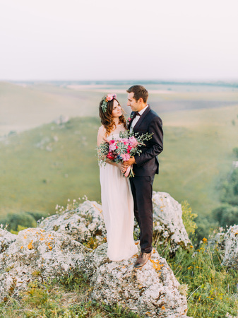 Newlywed couple is looking in the eyes of each other while standing on the rock at the background of the green mountains.の写真素材