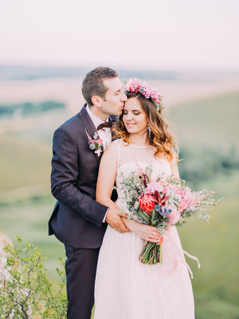 The groom is hugging the bride back while kissing on the forehead. Romantic portrait in the mountains.の写真素材