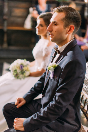 The half-length side portrait of the groom sitting on the chair during wedding ceremony in the church at the blurred background of the bride.の写真素材