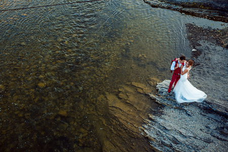 Cheerful stylish newlywed couple is hugging on the river bank during the sunny day. Above view.の写真素材