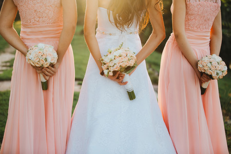 The bride and bridesmaids in an elegant dress is standing and holding hand bouquets of pastel pink flowers and greens with ribbon at nature. Young beautiful girls holds a wedding bouquet outdoors.の写真素材