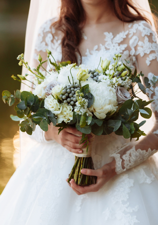 Young beautiful girl in an elegant dress is standing and holding hand bouquet of pastel flowers and greens with ribbon at nature. The bride holds a wedding bouquet outdoorsの写真素材