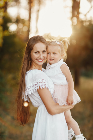 a woman holds a child in her arms in the park. young blonde mother holding her toddler daughter in her arms as they are walking through the park on a sunny day. little girl with two tailsの写真素材