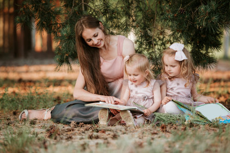Outdoor portrait of two little girl is reading a book on the grass with mother. She has a look of...