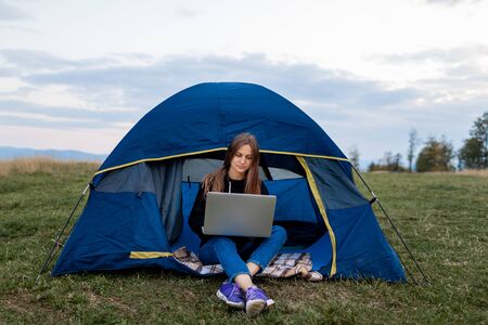 Girl with laptop in the mountains. Tourist woman uses a laptop in nature, freelancer typing on a computer near a tent in a journey against the backdrop of a mountain landscape.の写真素材