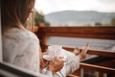 young woman in stylish nightwear enjoy drinking coffee or tea outdoor on balcony in the morning and looks at the mountains.の写真素材