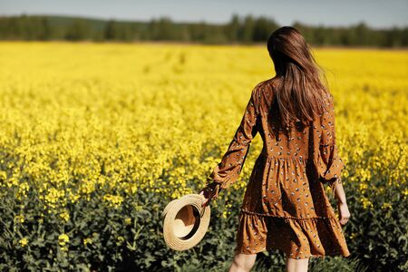 stylish young woman in a field of yellow flowers. Girl in straw hat and in a floral dress. place for inscription. rear view. background with yellow flowers and blue sky.の写真素材
