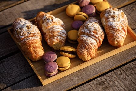 Freshly baked french croissants and colored macaroons or macarons on wooden tray . picnic on sunny day on wooden bridge near lake. Top view, close-upの写真素材
