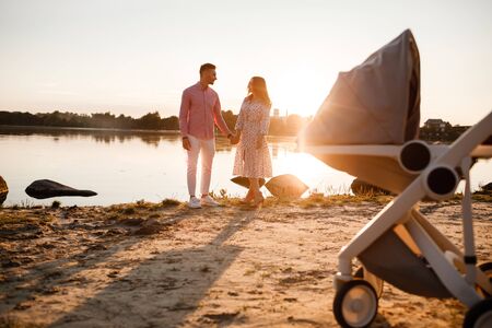 Beautiful young loving couple with baby walking on the lake at sunset. baby stroller on the front background. family has a good time together. copy space.の写真素材