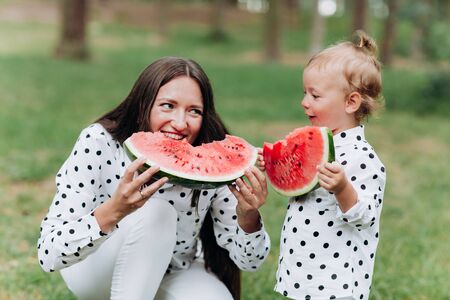 happy mother and daughter eat watermelon in summer park. Happy smiling family eating watermelon in park. Mother and daughter spend time together. Diet, vitamins, healthy food conceptの写真素材