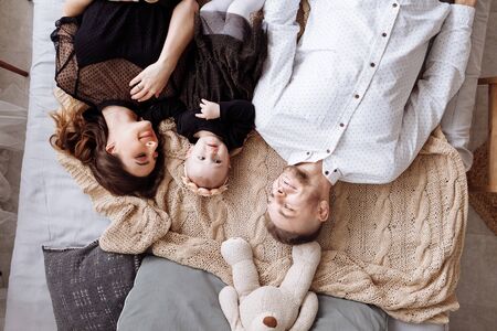 Beautiful young family lying together on the bed at home, mom, dad and baby girl, top view. Mother's, father's, baby's day. Family spending time together. Family look. selective focus.の写真素材