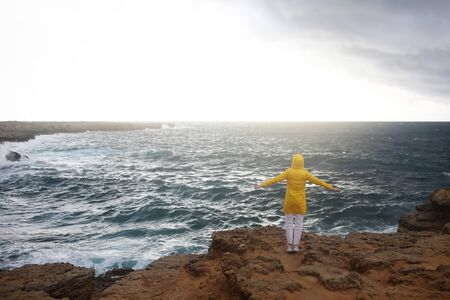 young woman dressed in yellow raincoat standing with outstretched arms while enjoying beautiful sea landscape in rainy day on the rock beach in cloudy spring weather.の写真素材