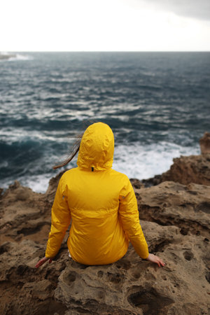 young woman dressed in yellow raincoat sitting on the cliff looking on big waves of the sea while...