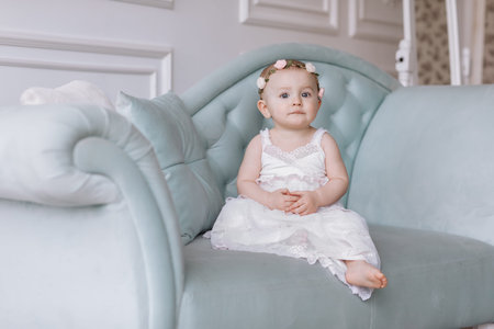 cute little girl in white dress and hoops of flowers on head is sitting on a couch in a classic style and having fun at home..の写真素材