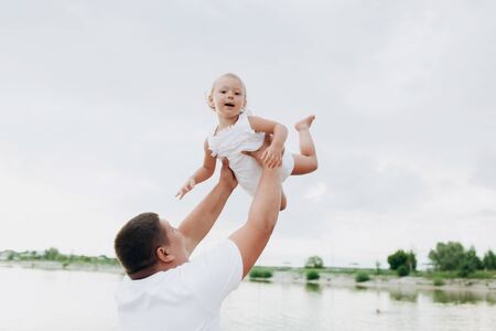 Father throws baby on pier on lake background, girl flies in the sky. Portrait dad with child together. Daddy, little daughter outdoors. Family fishing holiday on pondの写真素材