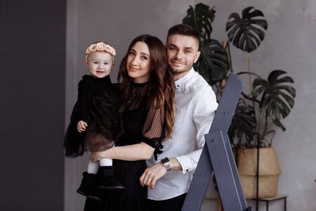 Portrait of beautiful young parents and their cute little daughter hugs, looking at camera and smiling indoors. mother's, father's, baby's day. concept of family. family lookの写真素材