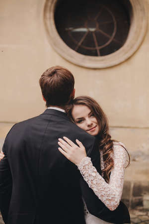 Wedding couple hugs near the vintage green door. Stone walls in ancient town background. bride with long hair in lace dress and groom in suit and bow tie. Tender embrace. Romantic loveの写真素材