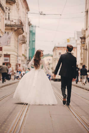 Happy newlyweds couple on a walk in old European town street, gorgeous bride in white wedding dress together with handsome groom. rear view.の写真素材