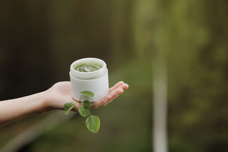 woman's hands holding jar of cosmetic aloe gel next to green leaves and branches on nature...