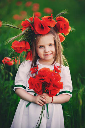 beautiful little girl with a wreath of poppies on head collects a bouquet of wildflowers. cute child in poppies field.の写真素材