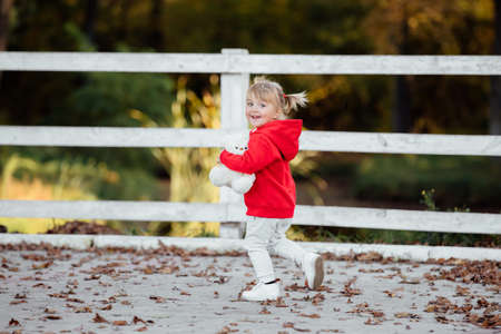 Little Cute Girl dressed in red sporty clothes in the autumn park, playing with a teddy bear on sunny day.の写真素材