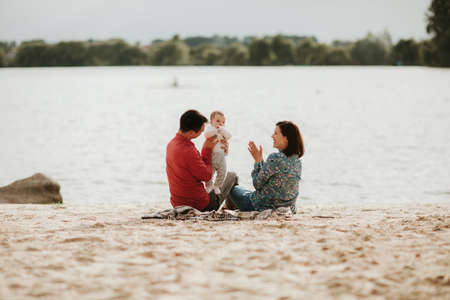 Happy young family sitting near the water. Mom, dad and their little baby daughter sitting together. Happy family outdoors. Smiling peopleの写真素材