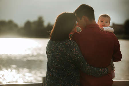 Happy young family sitting near the water. Mom, dad and their little baby daughter sitting together. Happy family outdoors. Smiling peopleの写真素材