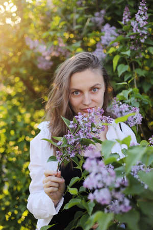 unreal stunning beautiful young woman walking in a green flowered in spring in a lavender garden with a bouquet of lilac in the hands. Closeup fashion romantic portrait. smelling the flowers.の写真素材