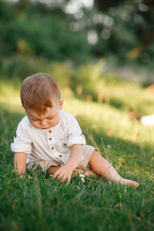 Happy adorable baby boy sitting on the grass in the park on summer day. Child in trendy and cute clothes.の写真素材