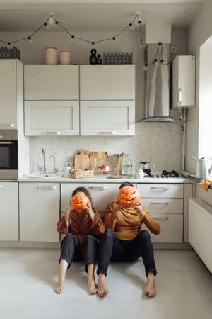 Young couple man and woman sitting on the floor at kitchen at home holding colorful pumpkins near their faces and preparing for halloween talking laughing cheerful.の写真素材