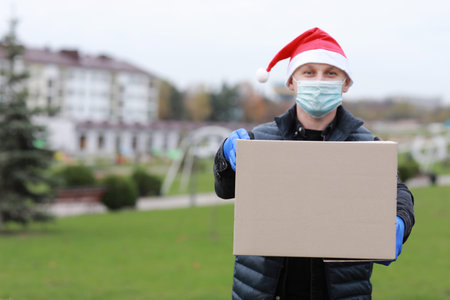 Delivery man in red cap, face medical mask and gloves hold paper bag near the car outdoor. Service...