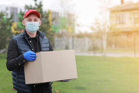 Delivery man in protective mask, gloves and santa's hat holding box in hands outdoors, delivery...
