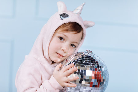 Smiling beautiful little girl in unicorn pink costume is smiling and having fun with disco ball on a blue wall background. happy childhoodの写真素材