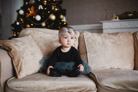 cute baby girl in dress and bow on head on bed with Christmas background behind