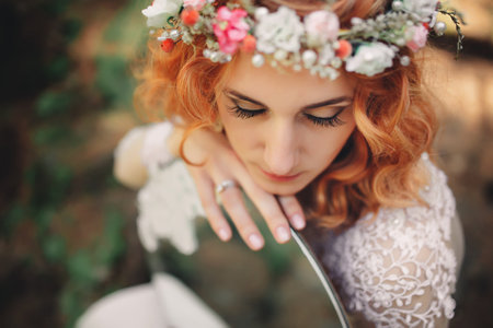 beautiful young red-haired bride in the forest with a floral wreath on her head and mirror. woman in long white dress outdoors on summer day. wedding day.の写真素材