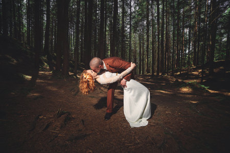 Happy stylish couple newlyweds in the green forest on summer day. bride in long white dress and groom in red suit are hugging. wedding dayの写真素材