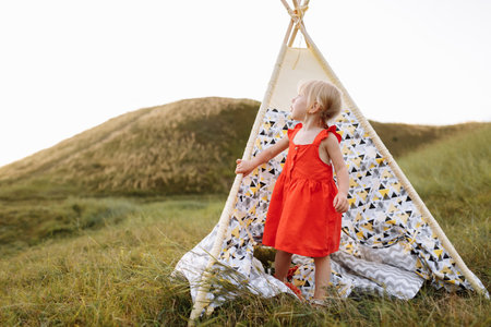 Portrait of a little beautiful girl in red dress on nature on summer day vacation. The playing in the green field at the sunset time. Close Up. The concept of family holiday and time togetherの写真素材