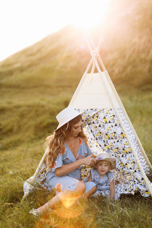 Beautiful mother with her little daughter near wigwam in the field. Spending time together, outside, on vacation, outdoors. Beautiful sunset light in the garden or in the parkの写真素材